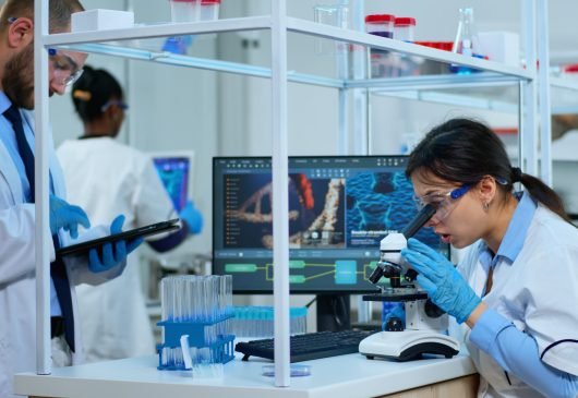 Scientist team checking virus development using microscope and writing analysis results on tablet in modern equipped lab. Multiethnic group examining vaccine evolution using high tech for research.
